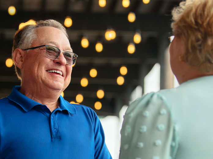Gray haired man in a blu shirt talking to a blonde haired woman in a mint green shirt under cafe lights.